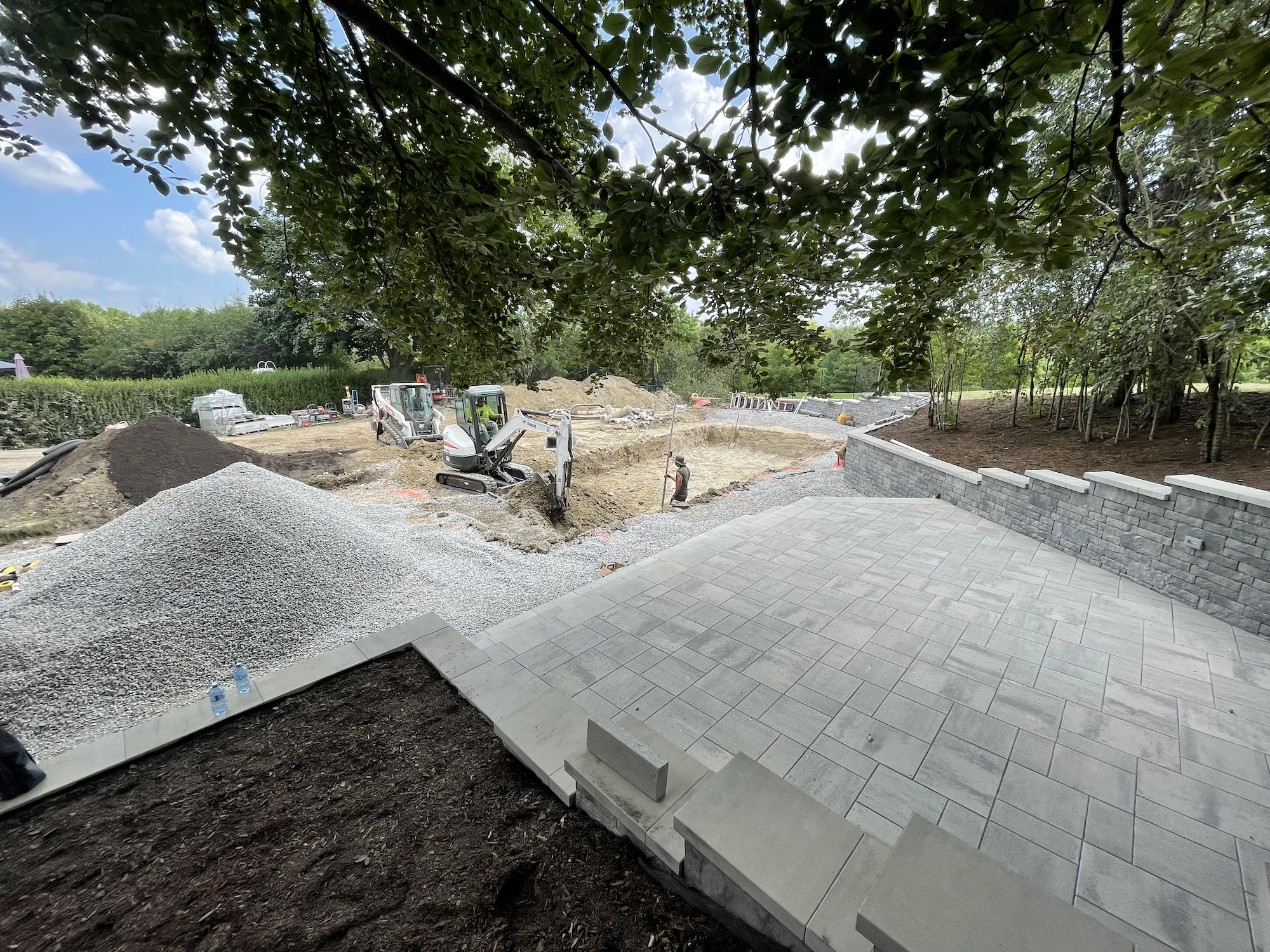 Construction site with paved area and groundwork, surrounded by trees and greenery.