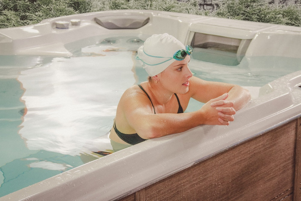 A swimmer resting on the edge of a Sundance® Surf™ 17 Swim Spa after a training session, with a wooden cabinet finish and serene backyard setting.