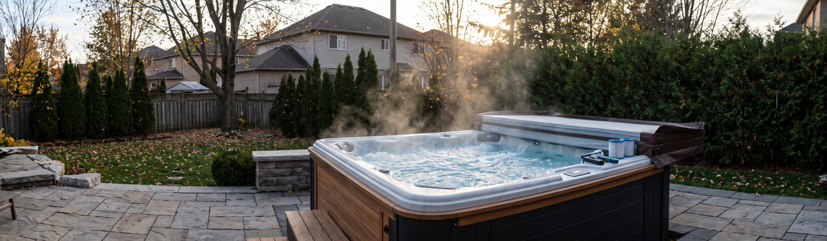 A steaming, bubbling outdoor hot tub on a natural stone patio in an Ajax, Ontario backyard during a cool late afternoon. A partially open winter cover and maintenance filters sit on the edge against a backdrop of suburban homes and evergreen privacy hedges.
