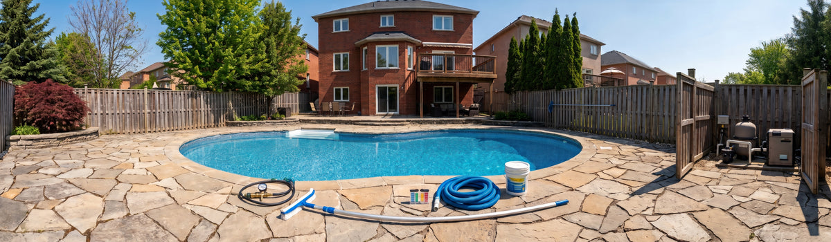 A backyard swimming pool in Ajax with a stone patio, wooden fence, brick house, trees, and pool cleaning equipment including a blue hose, brush, and bucket.
