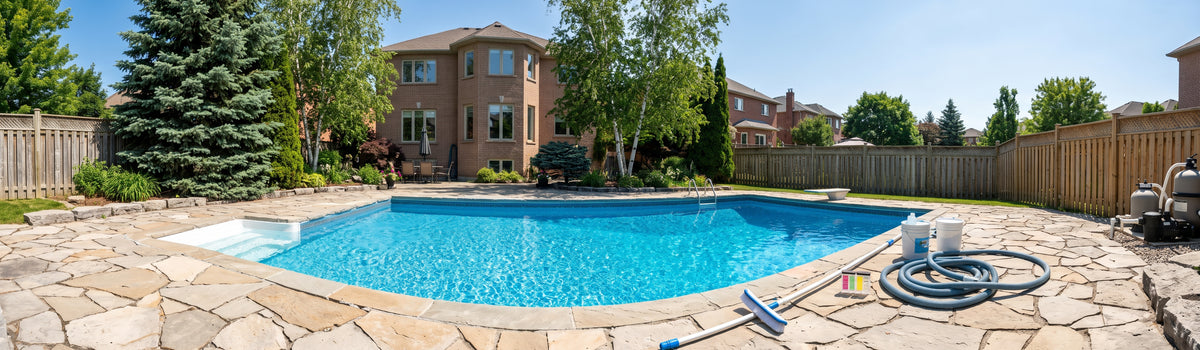 A backyard swimming pool with bright blue water, surrounded by a stone patio, wooden fence, trees, and a brick house under a clear blue sky.